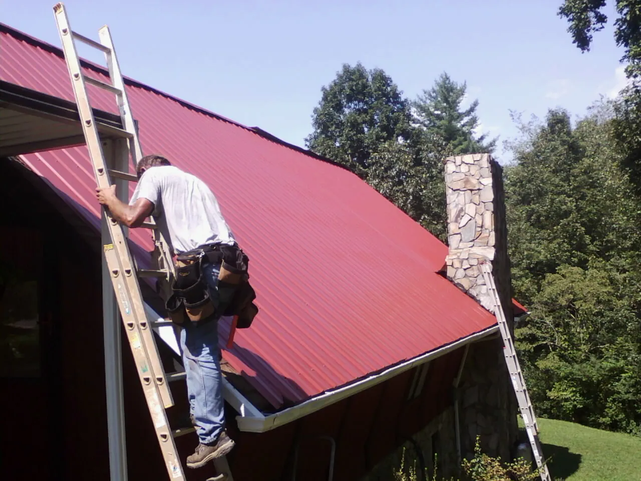 Roofer on a ladder beside a red metal roof and stone chimney — Soffit & Fascia Repair by Roofing Brothers in Minersville